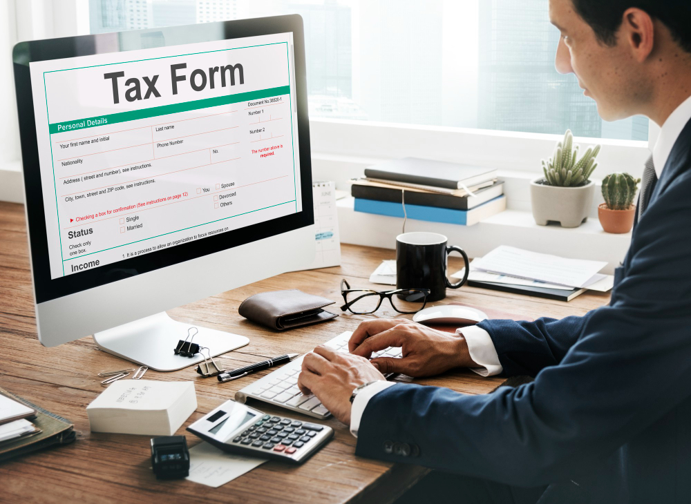 A businessman in a suit fills out a digital tax form on a desktop computer in a modern office, representing how AI-generated tax fraud schemes can exploit online filing systems and target unsuspecting professionals during tax season.