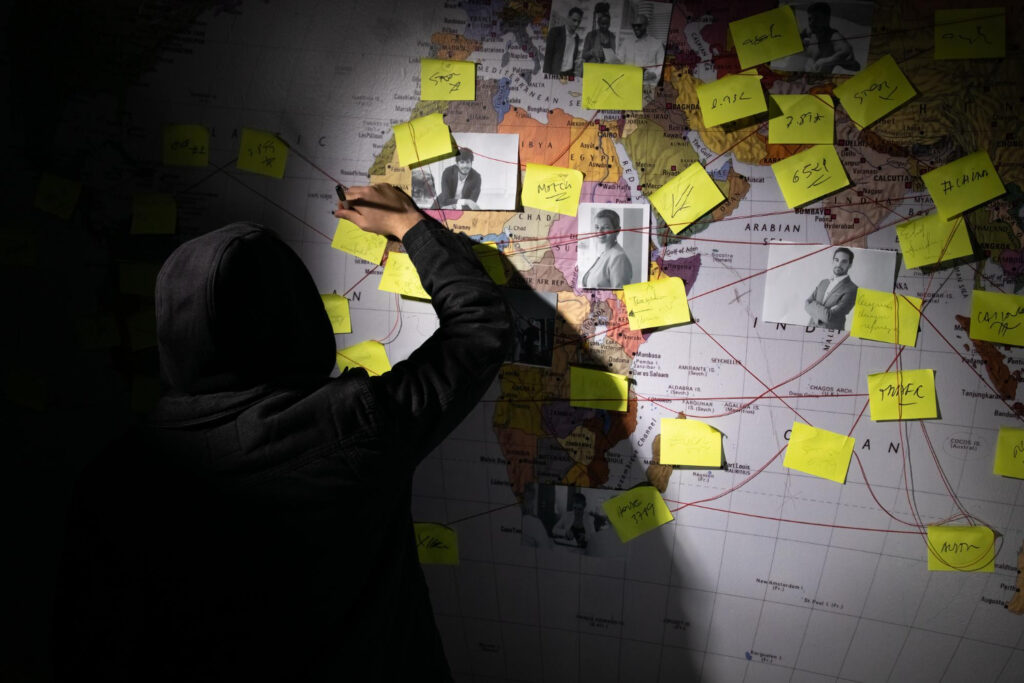 Back view of How Cybercriminals Target High-Net-Worth Individuals. Man planning hacker attack in dark room. Man writing on wall with stickers, photographs and red threads. Planning, conspiracy, hacking concept