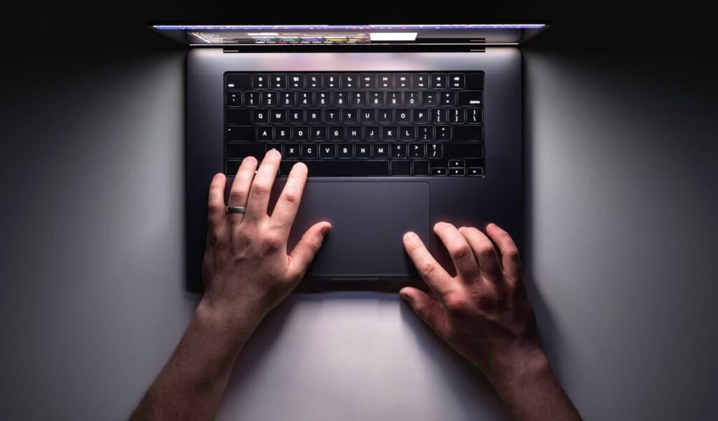 A person using a laptop in a dark environment with only the screen providing light, symbolizing cybersecurity vigilance—representing Dark Web Monitoring for Executives.